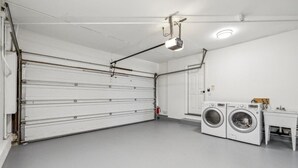 Une buanderie propre et blanche, dotée d’une grande porte de garage, de deux machines à laver et d’une petite table avec un évier.