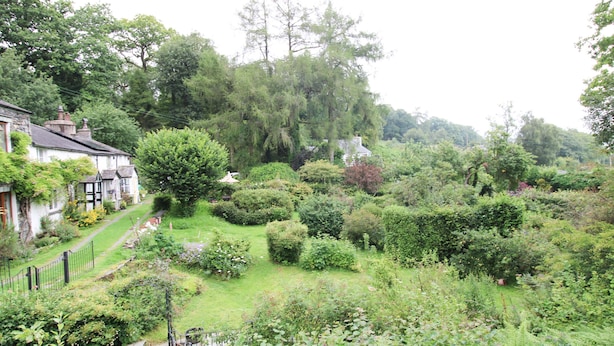 Ein üppiger Garten mit verschiedenen Pflanzen und Bäumen, ein Weg, der zu einem weißen Gebäude mit schwarzem Dach führt, und ein Zaun, der das Anwesen umgibt.