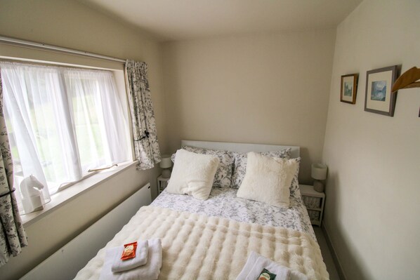 A bedroom with a bed, a window with curtains, a bedside table, and a wall with framed pictures. - Milldam Cottage, Coniston - a cottage that sleeps 4 guests in 2 bedrooms (Coniston)