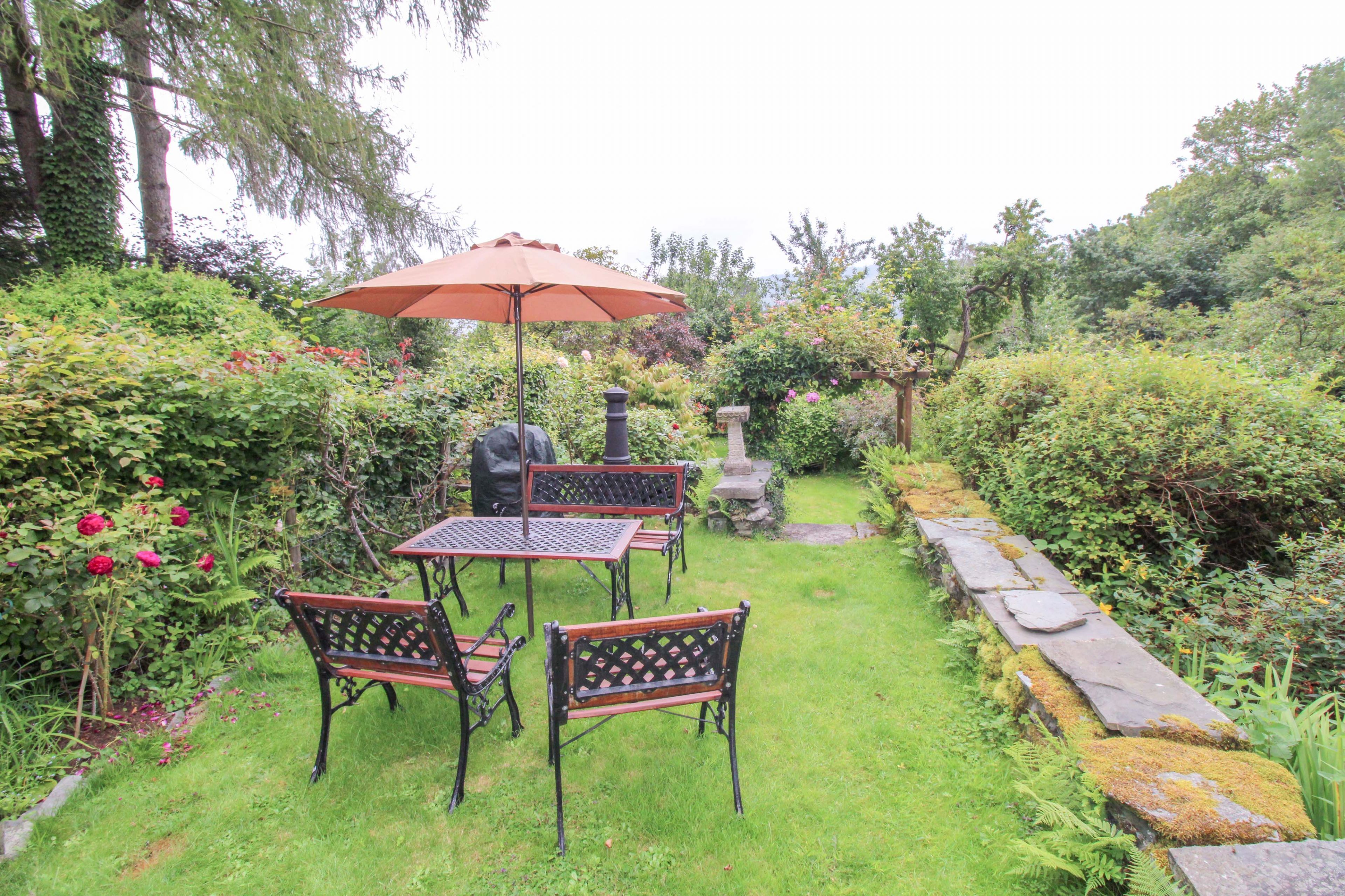 Een tuinpatio met een tafel en stoelen, een stenen pad en diverse planten en bloemen.