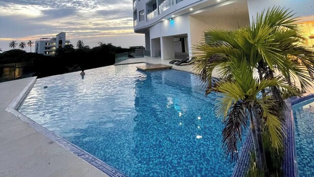 A modern hotel with a rooftop infinity pool, surrounded by palm trees and a clear sky.