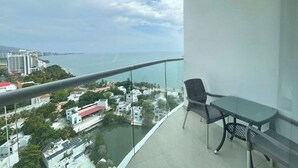 A balcony with a glass railing overlooking a coastal area, a table, and a chair.