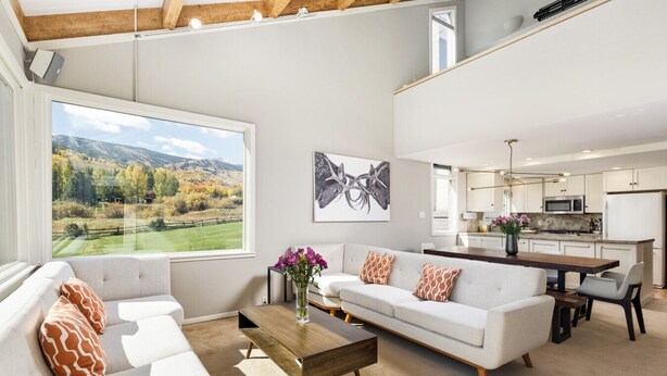 A modern living room with a large window, a sofa set, a coffee table, and a kitchen area in the background.