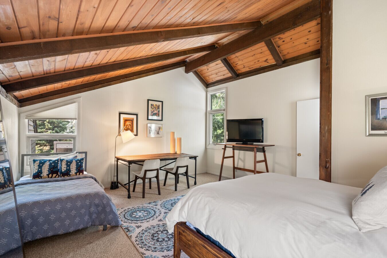 A bedroom with a sloped ceiling, a bed, a desk with chairs, a TV, and artwork on the walls.