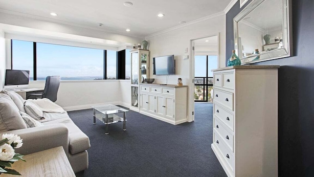 A modern living room with a sofa, coffee table, and a TV. There is a kitchen area with cabinets and a dining table. The room has large windows offering a view of the sky. - Awesome 1BR Apartment Kitchen Tennis Gym Hot Tub (Auckland)