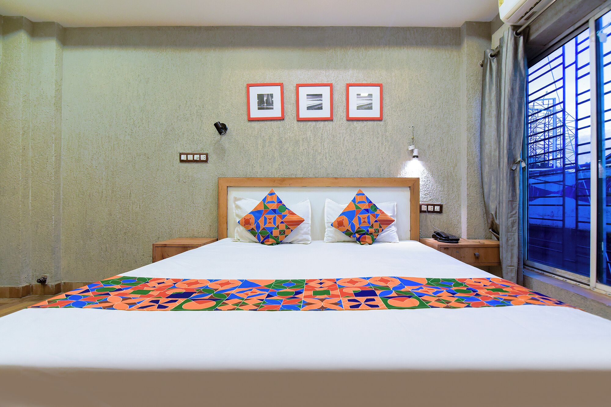 A hotel room with a bed, a nightstand, a lamp, and a window with a view of a building.
