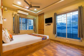 A hotel room with a bed, a ceiling fan, an air conditioning unit, a TV, and large windows with curtains.