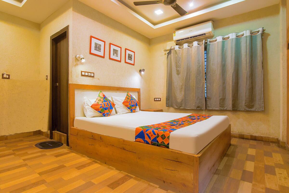 A hotel room with a wooden bed frame, a ceiling fan, an air conditioning unit, and a window with curtains.
