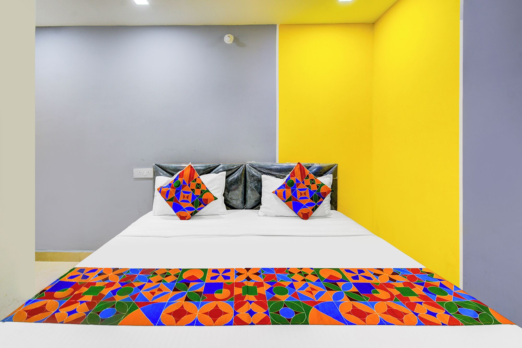 A bed with a colorful geometric-patterned bedspread and two pillows with a similar design, set against a wall with a grey and yellow color scheme.