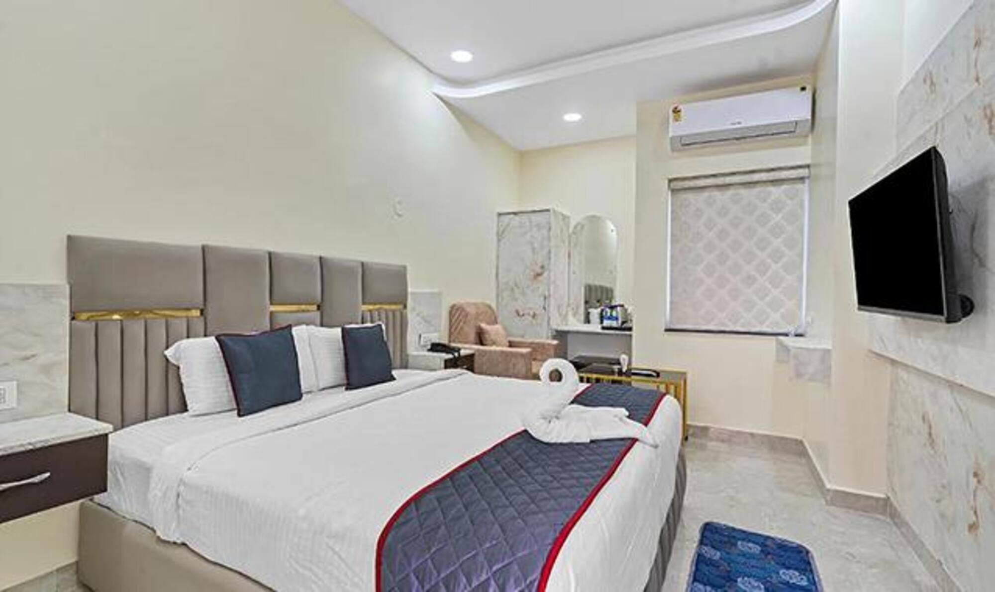 A hotel room with a large bed, a bedside table, a chair, a TV, and an air conditioning unit.