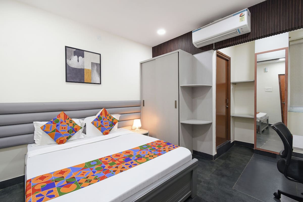 A hotel room with a bed, a bedside table, a lamp, an air conditioner, a wardrobe, a mirror, and a chair.