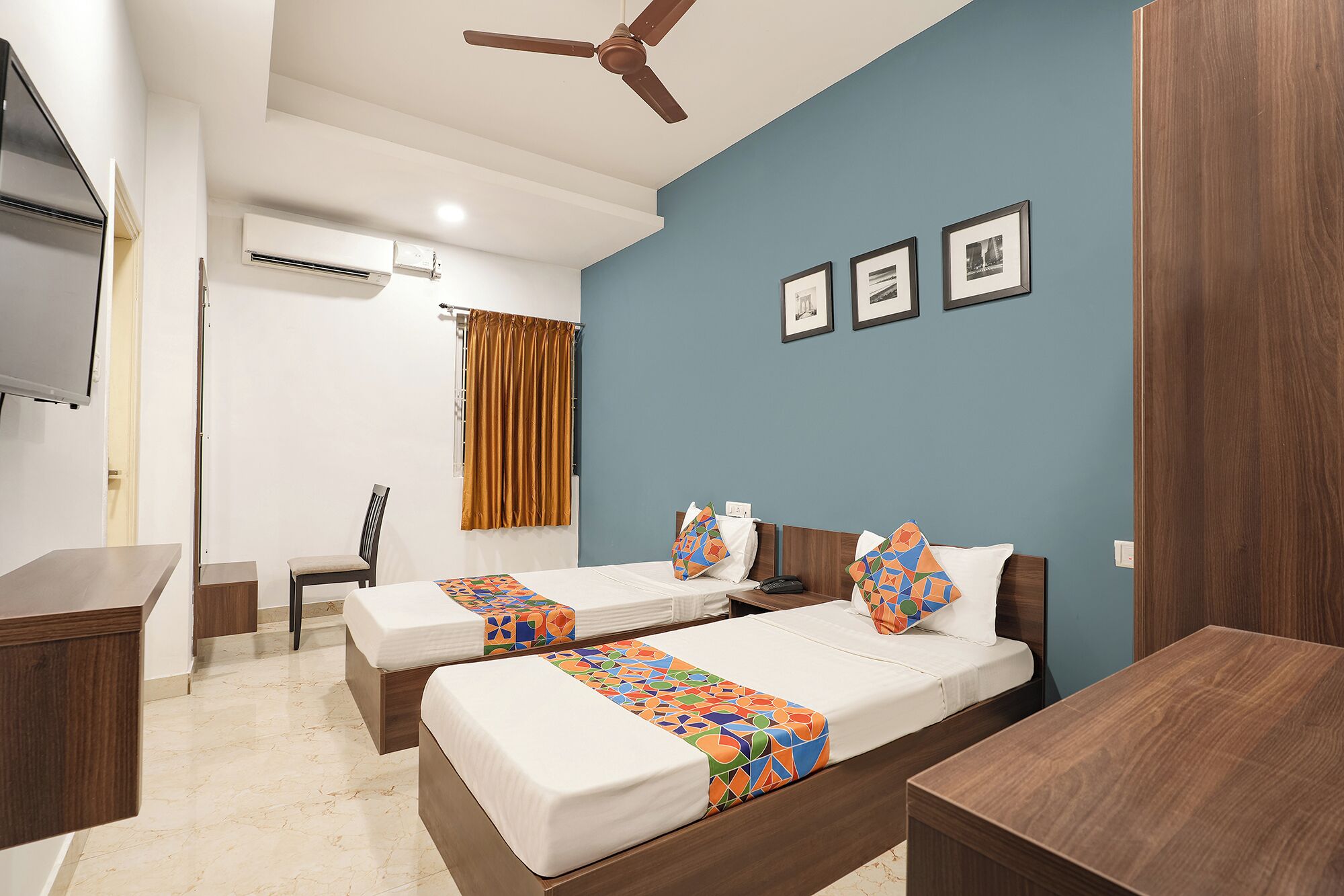 Habitación de hotel con dos camas, un escritorio, una silla, ventilador de techo y cuadros en la pared.