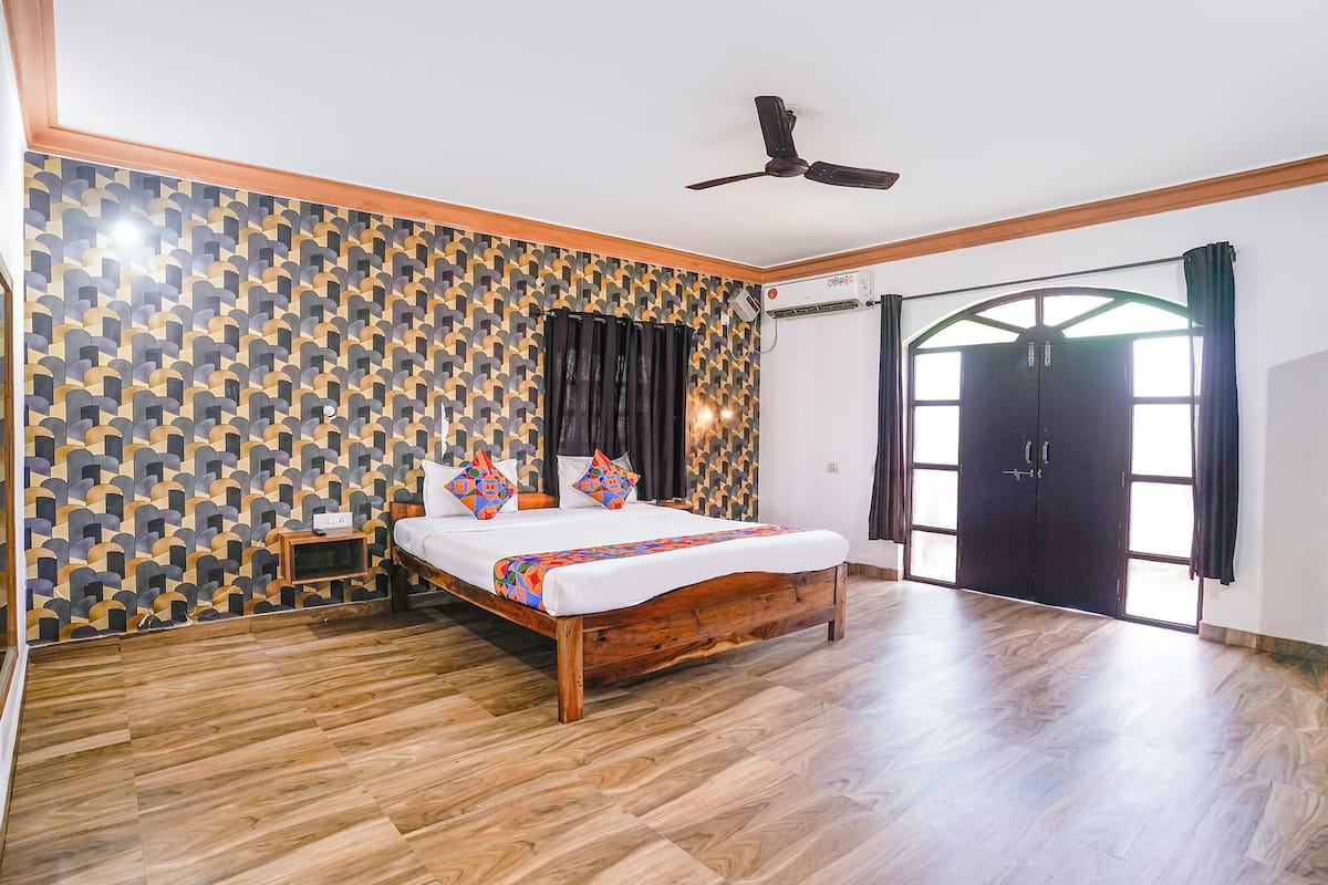 A bedroom with a bed, wooden floor, geometric patterned wallpaper, a ceiling fan, and a large window with curtains.