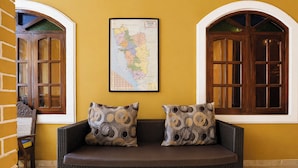 A wicker sofa with patterned cushions, a framed map on the wall, and wooden-framed windows. - Via Romeo's Stay - A Beach Property (Calangute)