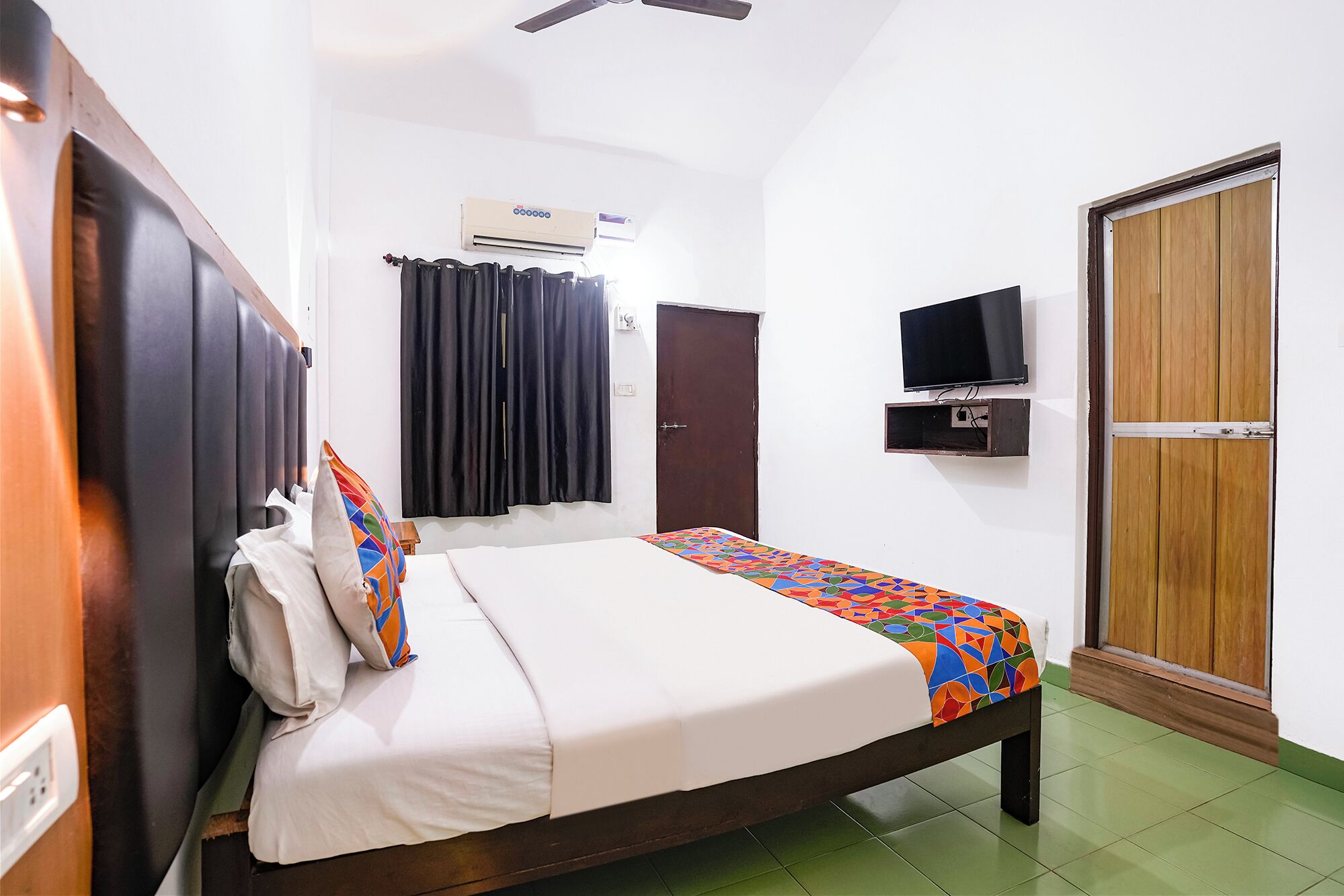 A single bed with a colorful geometric-patterned bedspread, a television mounted on the wall, and a wooden wardrobe.