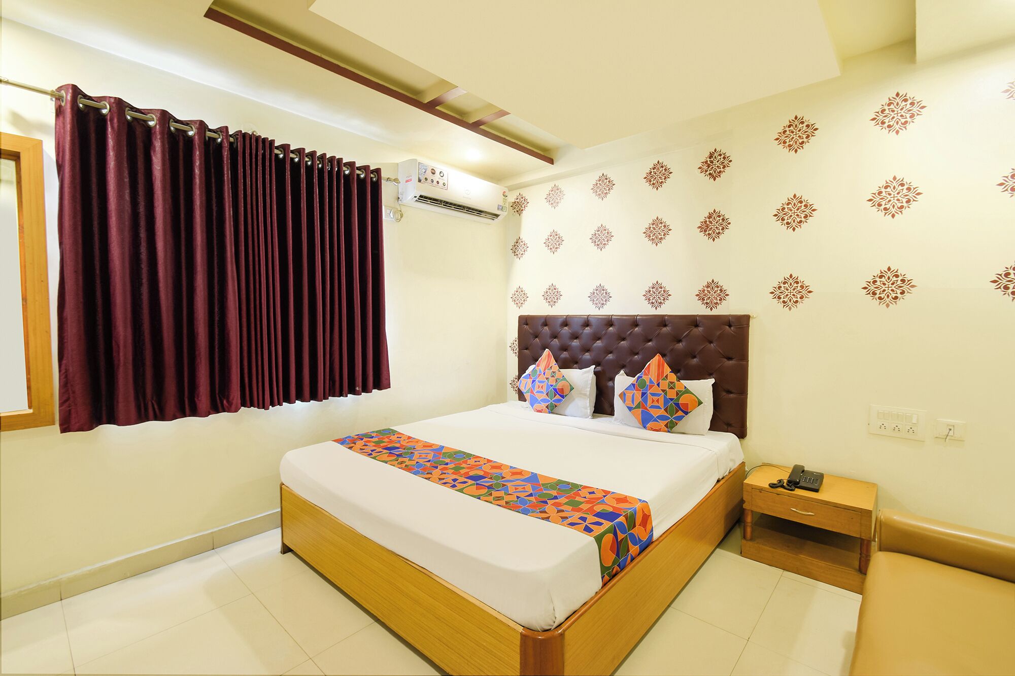 A neatly arranged hotel room with a bed, a bedside table with a phone, and a wall-mounted air conditioning unit.