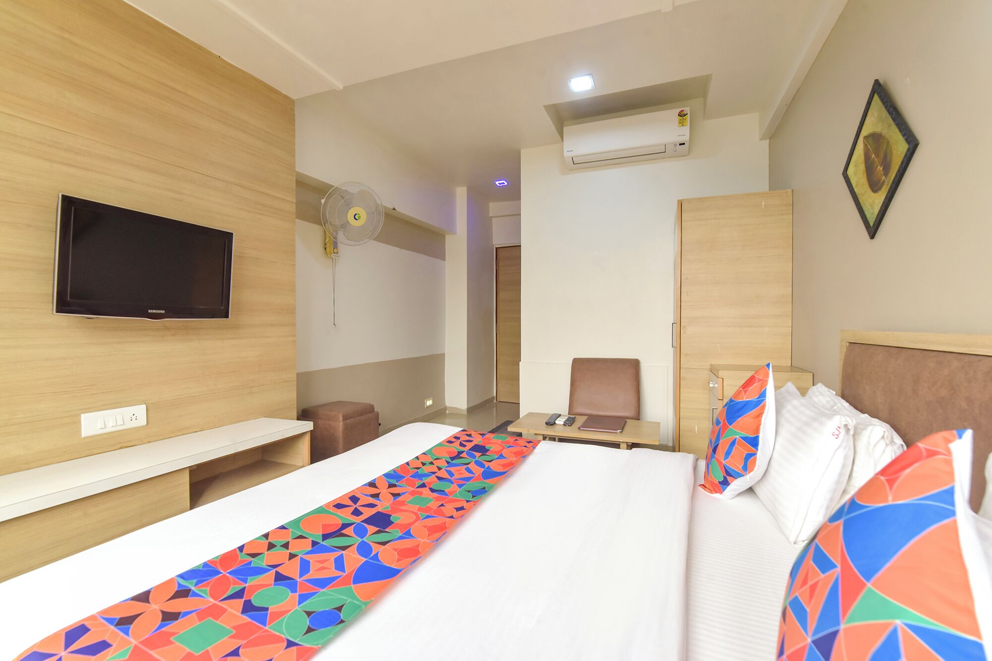 A hotel room with a bed, a TV, a desk, a chair, and an air conditioning unit.