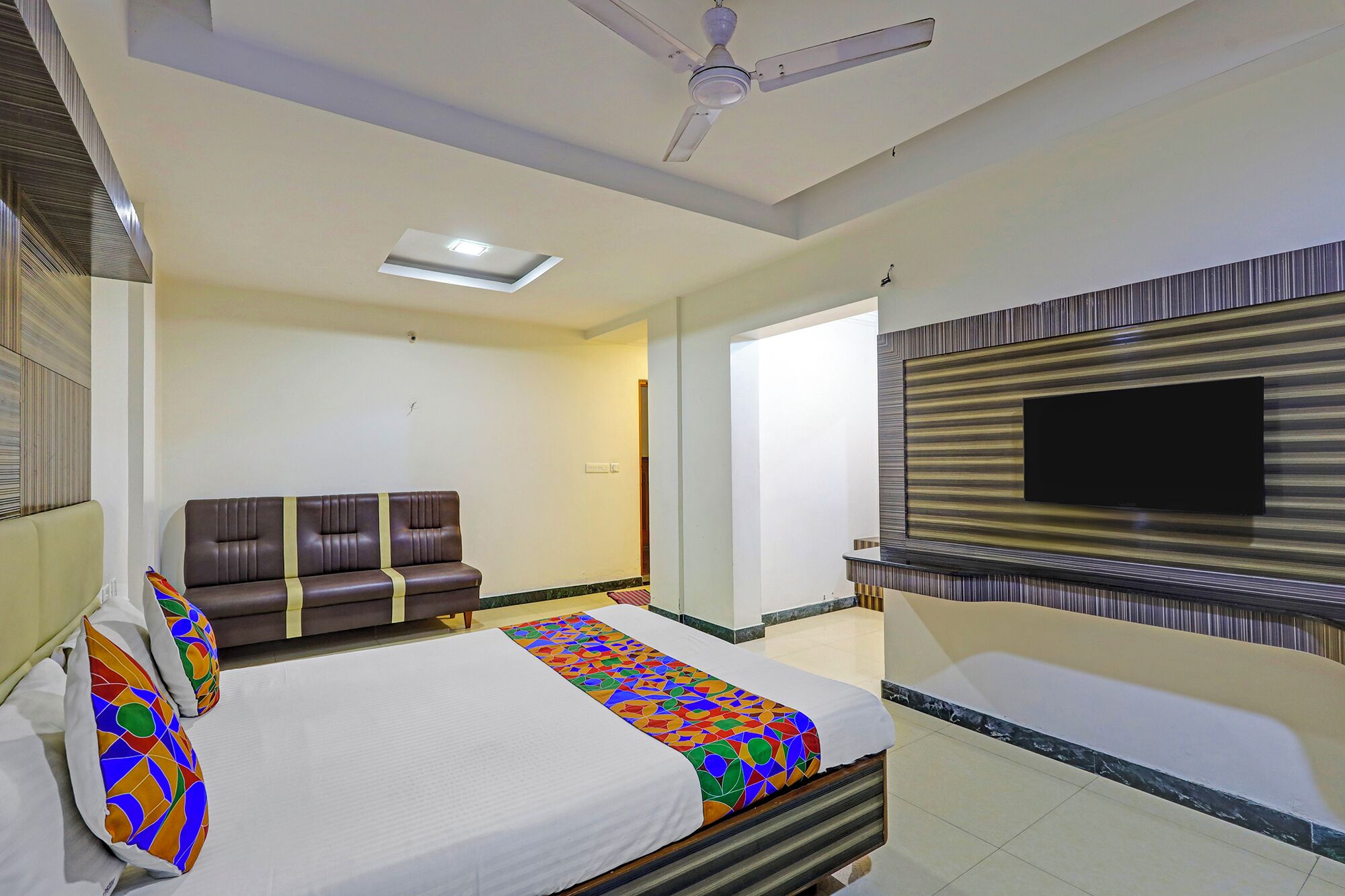 A hotel room with a bed, a sofa, a ceiling fan, a TV mounted on the wall, and a patterned rug.