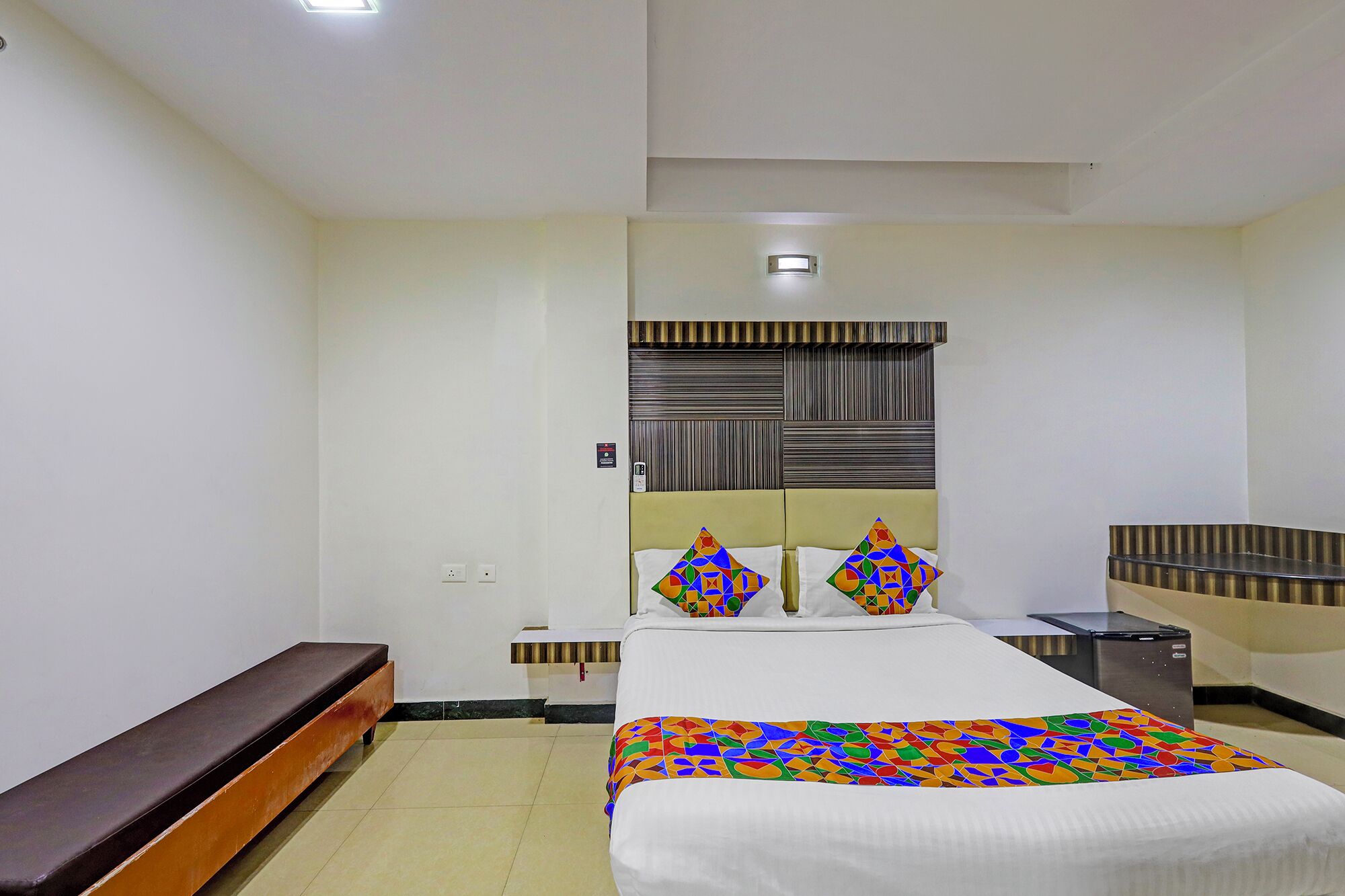 Una habitación de hotel con cama, mesita de noche, banca y un pequeño escritorio con lámpara.