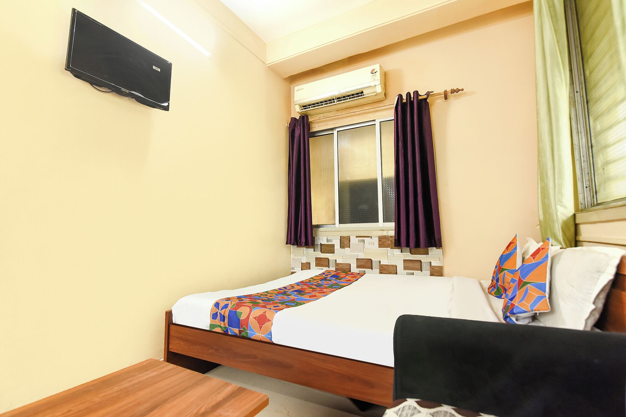 Una habitación de hotel con cama, mesita de noche de madera, escritorio, unidad de aire acondicionado y ventana con cortinas.