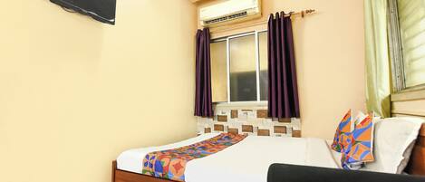 Una habitación de hotel con cama, mesita de noche de madera, escritorio, unidad de aire acondicionado y ventana con cortinas.