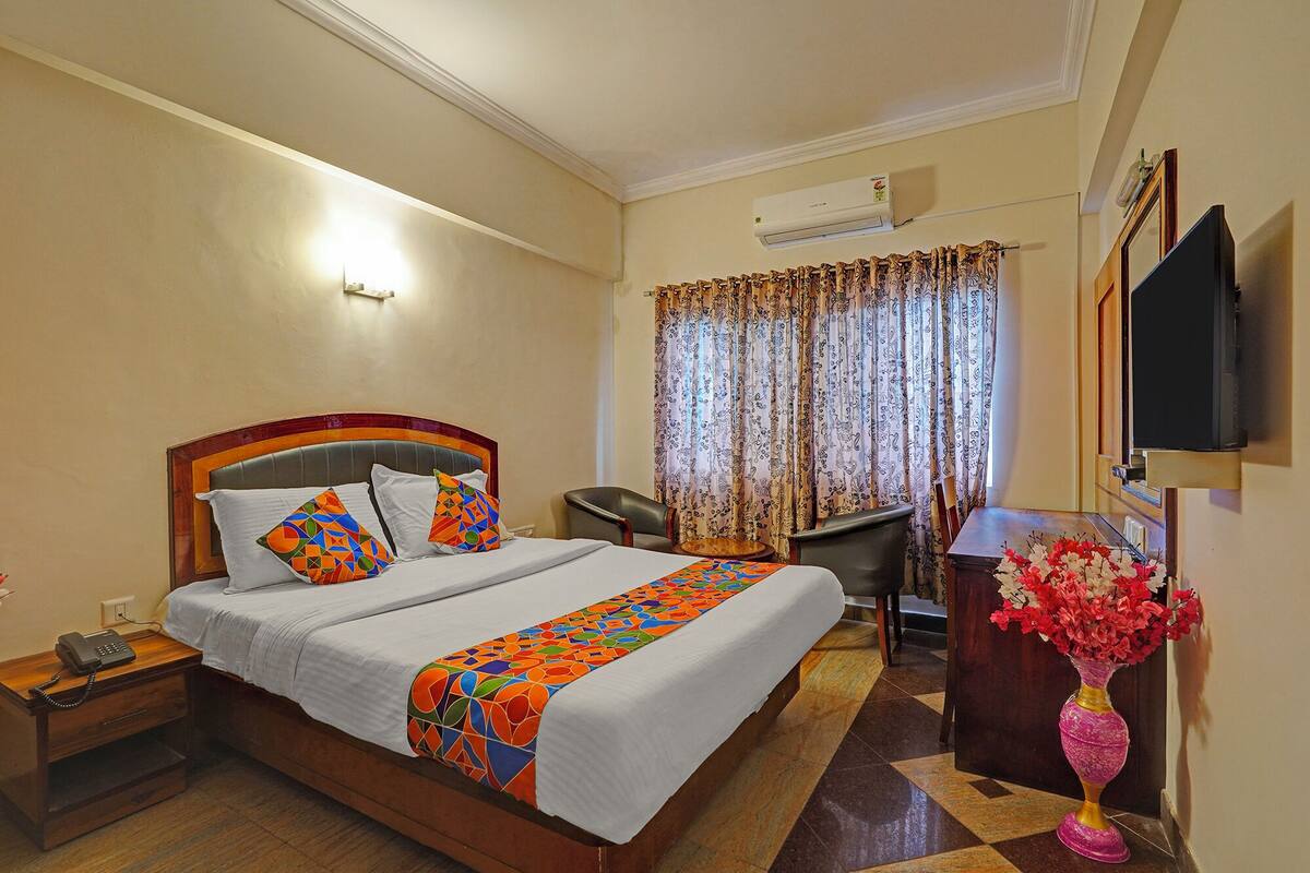 A hotel room with a bed, bedside table, chair, desk, TV, and a vase with flowers.