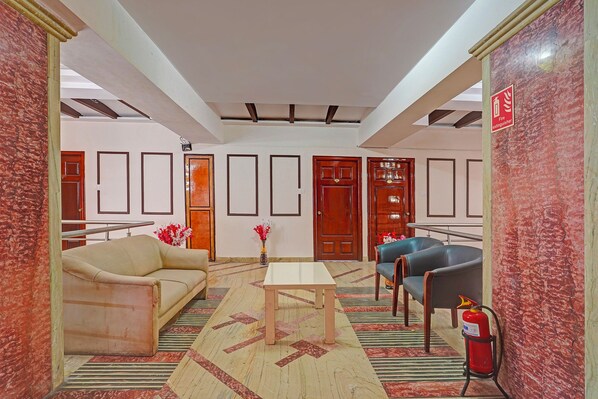 A hotel lobby with a beige sofa, a wooden table, and two blue chairs. There are framed pictures on the walls, a fire extinguisher, and a door with a wooden frame.