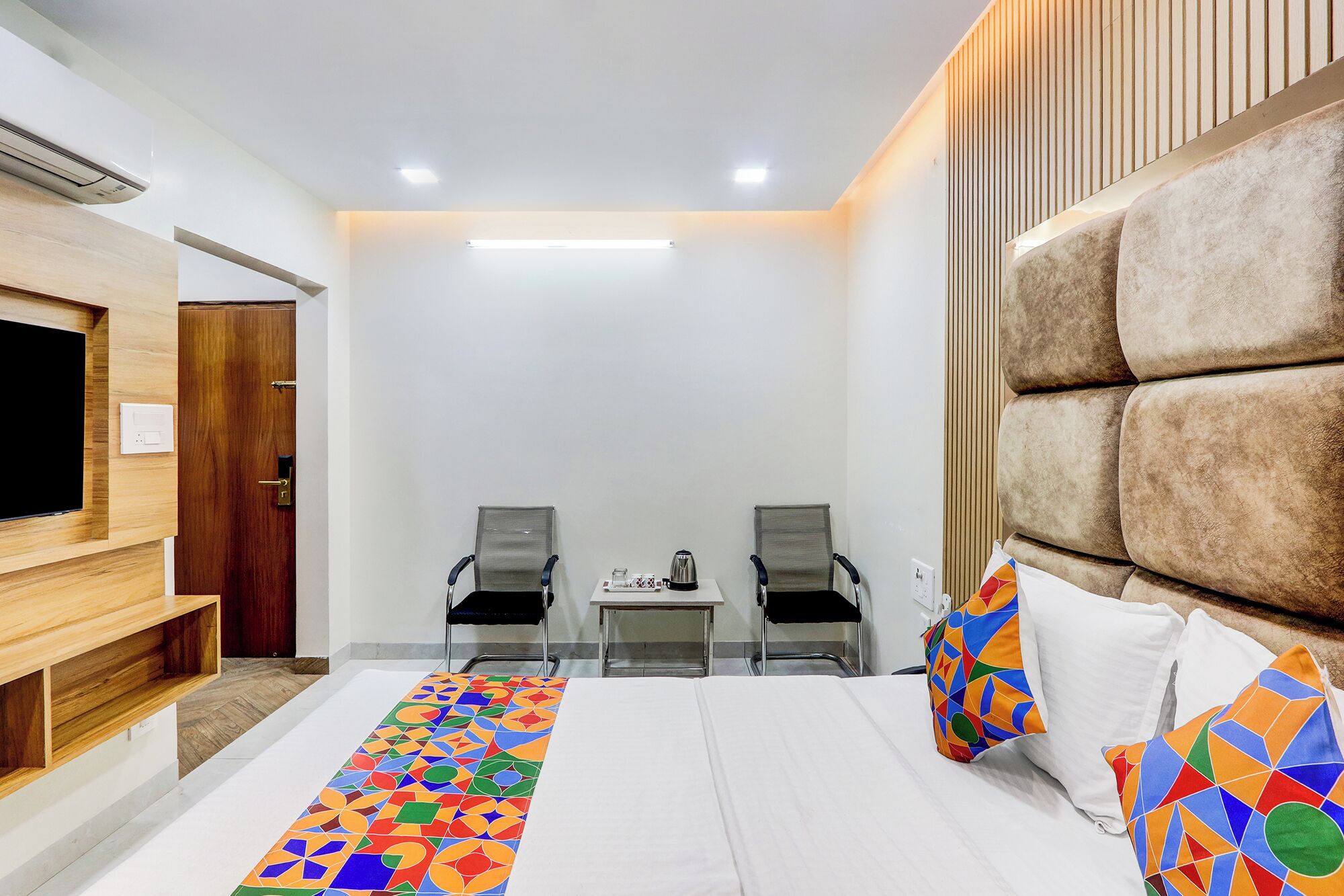 Habitación de hotel con cama, dos sillas, una mesita, televisión y un gran cabecero acolchado.
