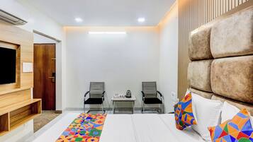 Habitación de hotel con cama, dos sillas, una mesita, televisión y un gran cabecero acolchado.