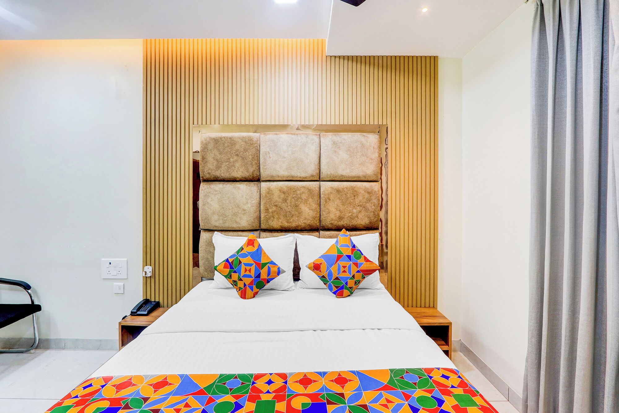 A modern hotel room with a large bed, a wooden headboard, and a colorful geometric-patterned bedspread.