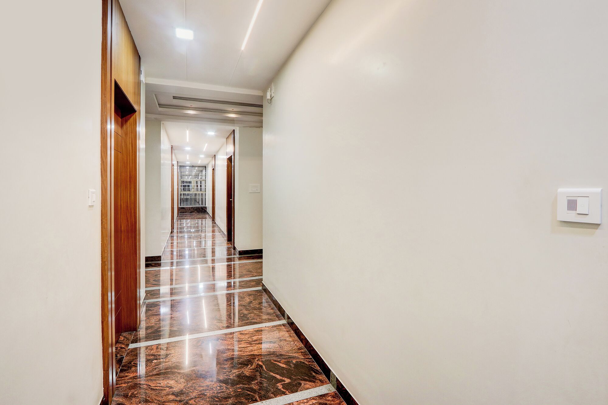Um corredor comprido e estreito com piso de mármore polido, paredes brancas e uma porta de madeira à esquerda.