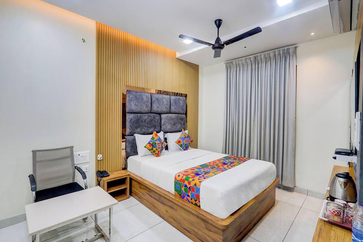 Une chambre d’hôtel moderne avec une tête de lit en bois, un lit agrémenté d’une literie aux motifs géométriques et colorés, un bureau avec une chaise, une petite table, un ventilateur de plafond et une fenêtre avec des rideaux.