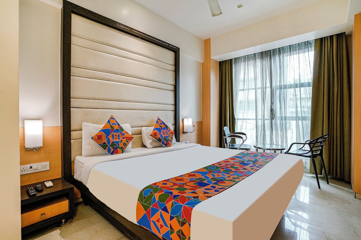 A hotel room with a large bed, a bedside table with a lamp, a chair, and a window with curtains.