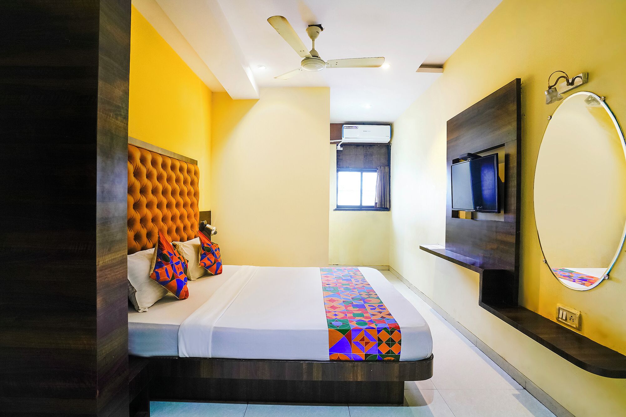 A hotel room with a tufted headboard, a bed with colorful bedding, a ceiling fan, a mounted TV, and a window with curtains.