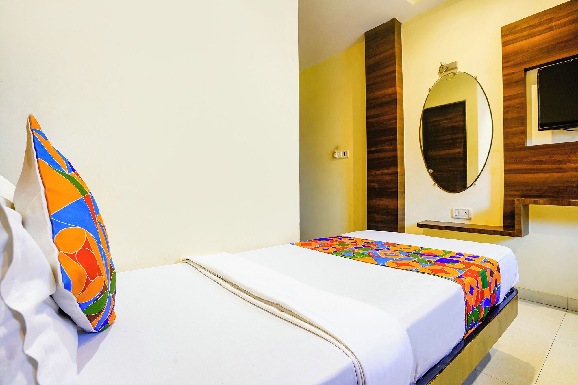 A hotel room with a bed, a colorful pillow, a mirror, and a TV.