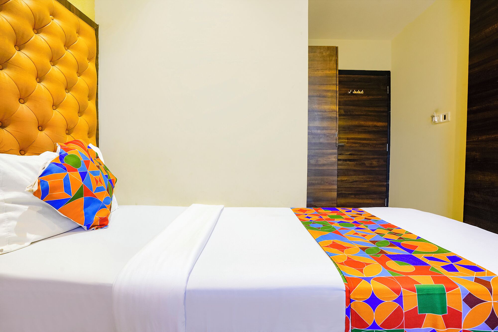 A hotel room with a tufted headboard, a colorful geometric-patterned bedspread, and a plain white bed.