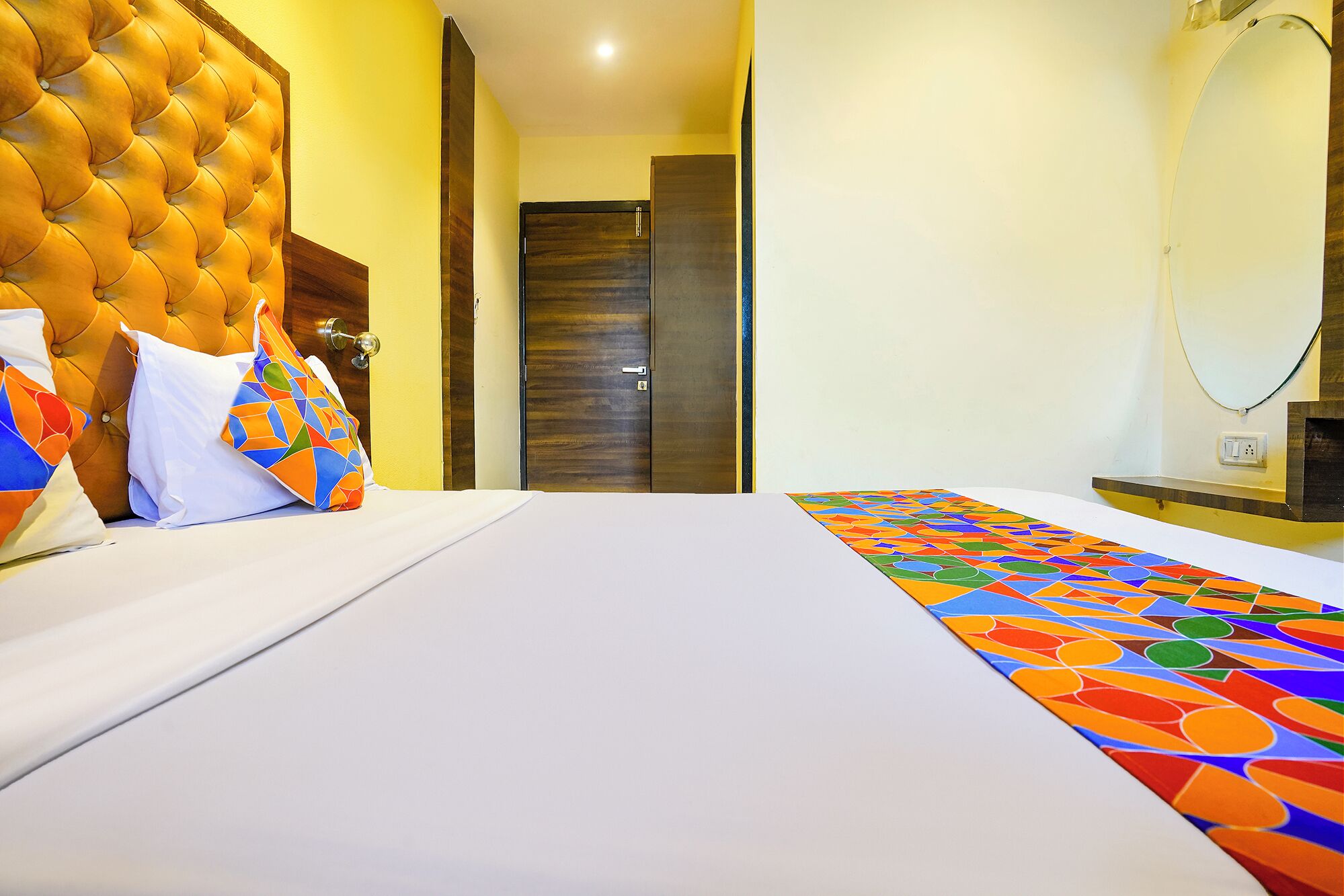 A hotel room with a tufted headboard, a bed with white linens, colorful pillows, and a patterned runner.