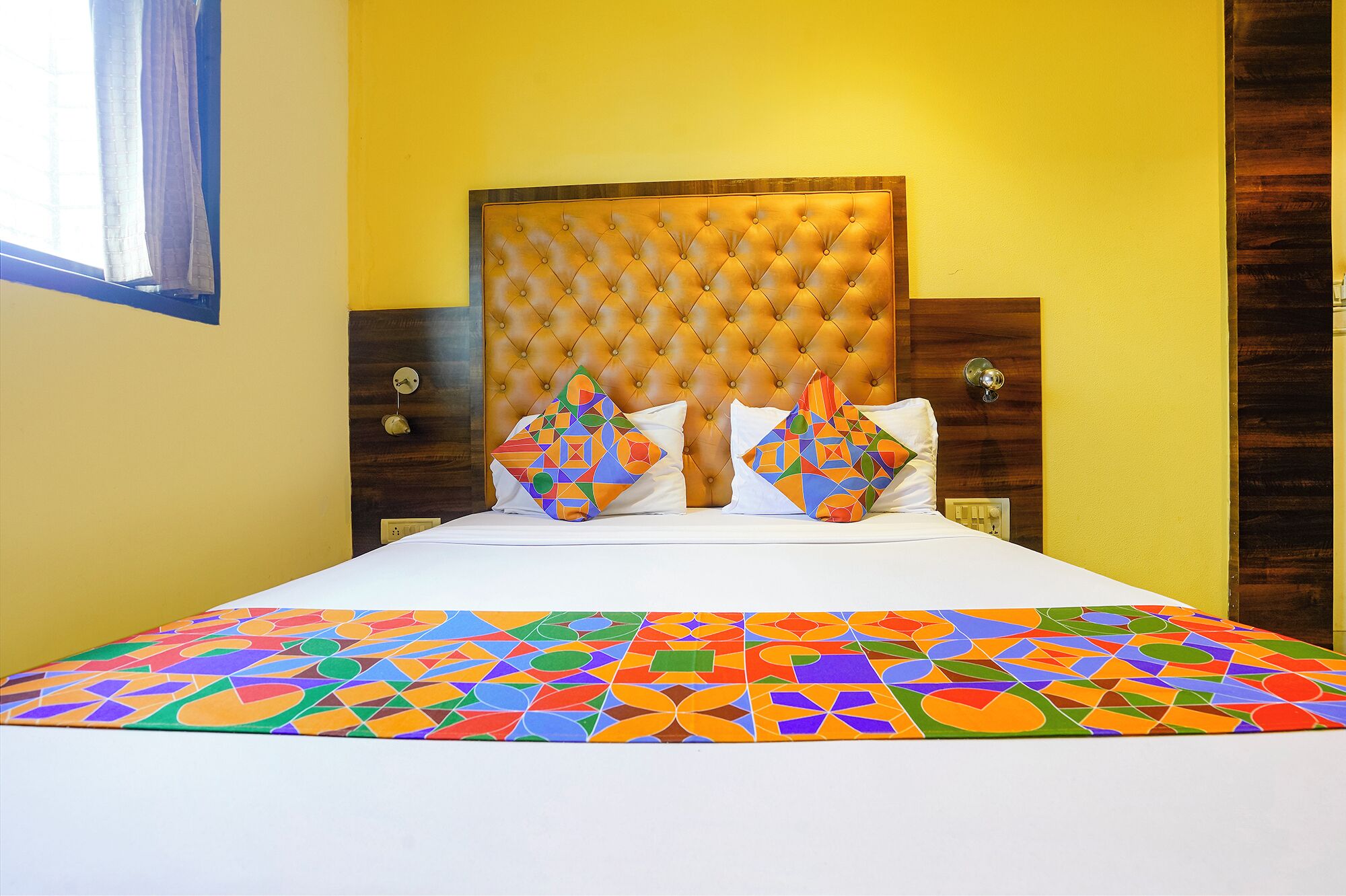 A hotel room with a tufted headboard, two pillows with geometric patterns, and a colorful bedspread.