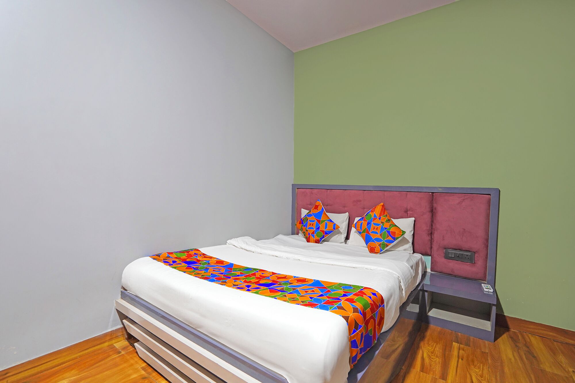 A bed with a colorful geometric-patterned bedspread, a white pillow, and a matching pillow on a headboard with a power outlet.