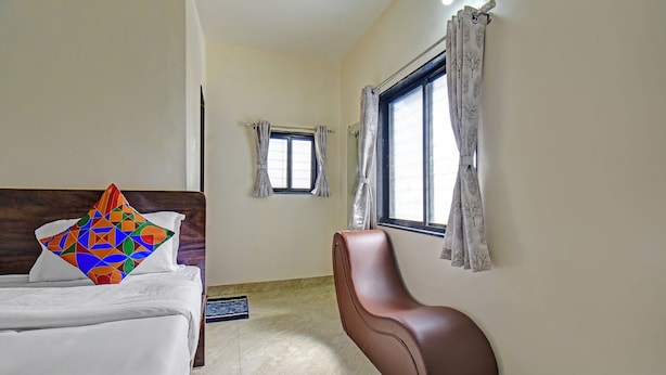 A hotel room with a bed, a brown chair, a window with curtains, and a small window with a view of the outside. - Fabhotel Shree Sai Inn (Paud)