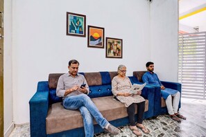 A blue sofa with three people seated, a wall with framed artwork, and a window with blinds. - FabHotel Lotus Prime Stay - Hinjawadi (Paud)