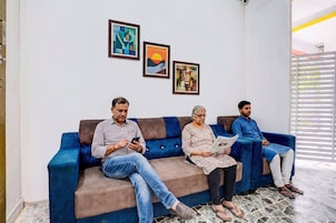 Un sofá azul con tres personas sentadas, una pared con obras de arte enmarcadas y una ventana con persianas.