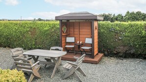 A wooden gazebo with a table and chairs, surrounded by a gravel area and a hedge.