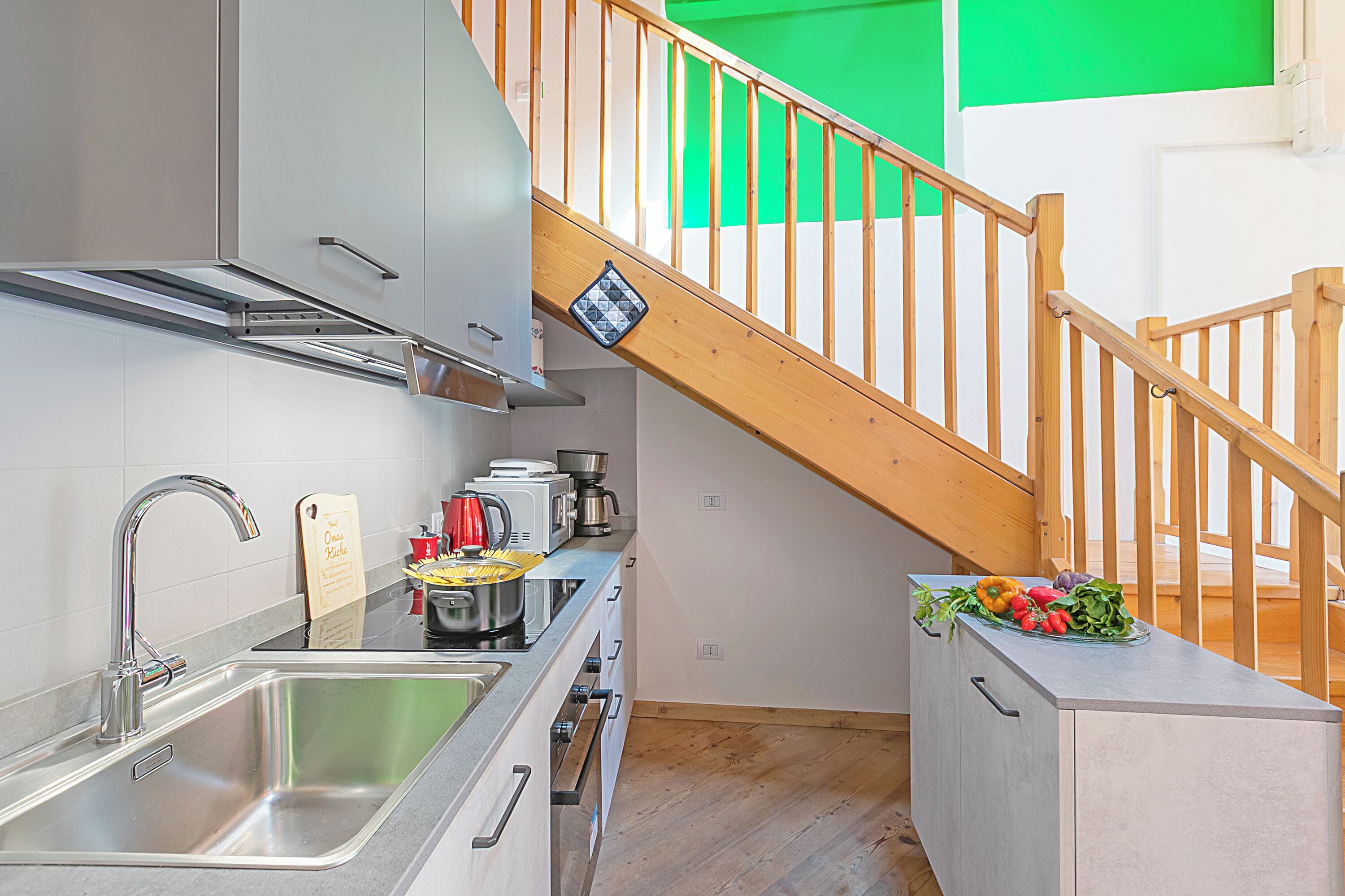 A modern kitchen with stainless steel appliances, a wooden staircase, and a small countertop with fresh vegetables.