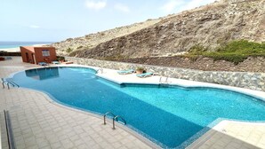 A pool with a stone wall and lounge chairs, a small building, and a rocky hillside in the background. - Welooveyou Top Victoria & Serenity (Costa Calma (incl. Urbanización Cañada del Río))