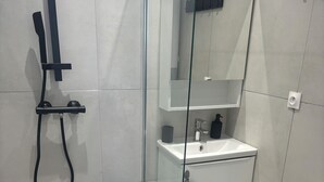 Et moderne badeværelse med et brusekabine i glas, en hvid vask med et spejl over, og et håndklæde hængende på væggen.