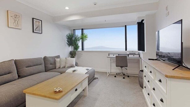 A modern, well-lit room with a grey sofa, a wooden coffee table, a desk with a chair, a television, and a large window offering a view of the sky and mountains. - Stunning Views of Rangitoto Kitchen Gym Rooftop Pool Parking (Auckland)