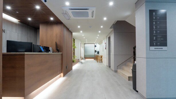 Un área de recepción moderna de hotel, con un mostrador de madera, una pantalla digital y una escalera que conduce a los pisos superiores.