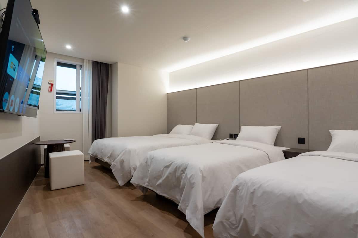 Ein Hotelzimmer mit drei Betten, einem Fernseher, einem kleinen Tisch und einem Fenster mit Vorhängen.