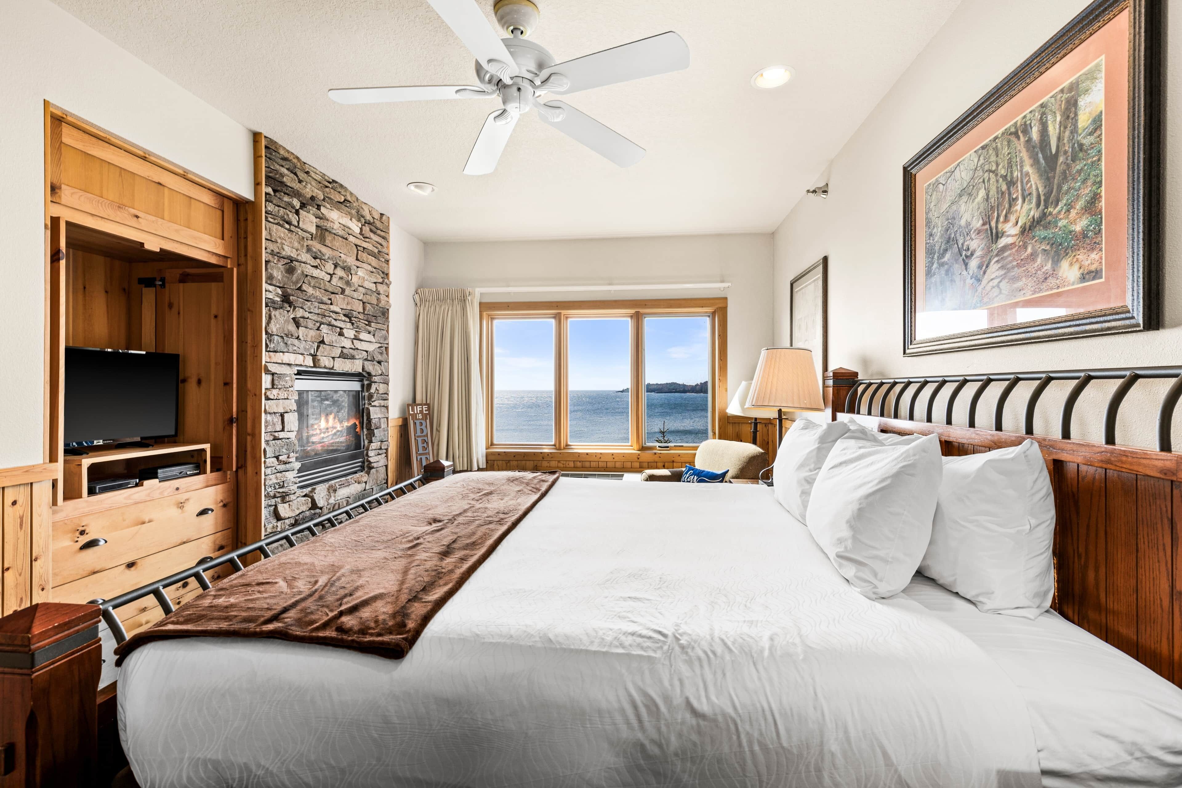 A hotel room with a large bed, a television, a stone fireplace, a view of the sea, and a ceiling fan.